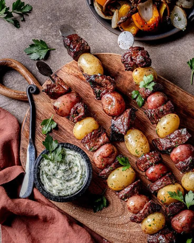 meat and potatoes skewered on a wooden cutting board next to a bowl of sauce