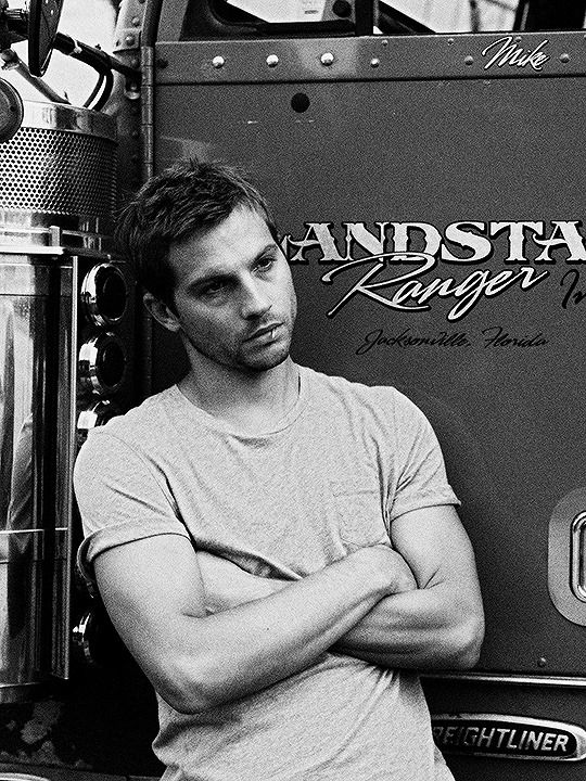 black and white photograph of a man leaning against the back of a firetruck