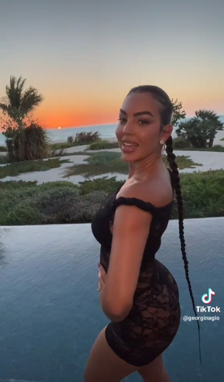 a woman standing next to a swimming pool in front of the ocean at sunset with her hands on her hips