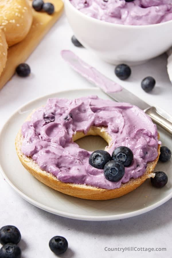 a bagel with blueberries and cream on it sitting on a plate next to some rolls