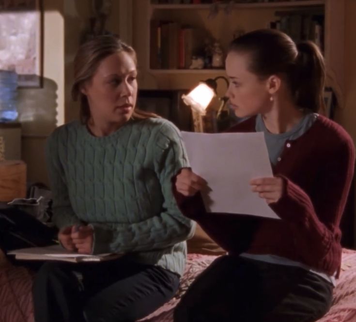 two women sitting on top of a bed talking to each other and holding papers in their hands