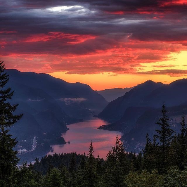 the sun is setting over some mountains with trees and water in the foreground, as seen from an overlook point