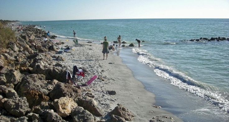 people are standing on the beach near the water and rocks, while others walk along the shore