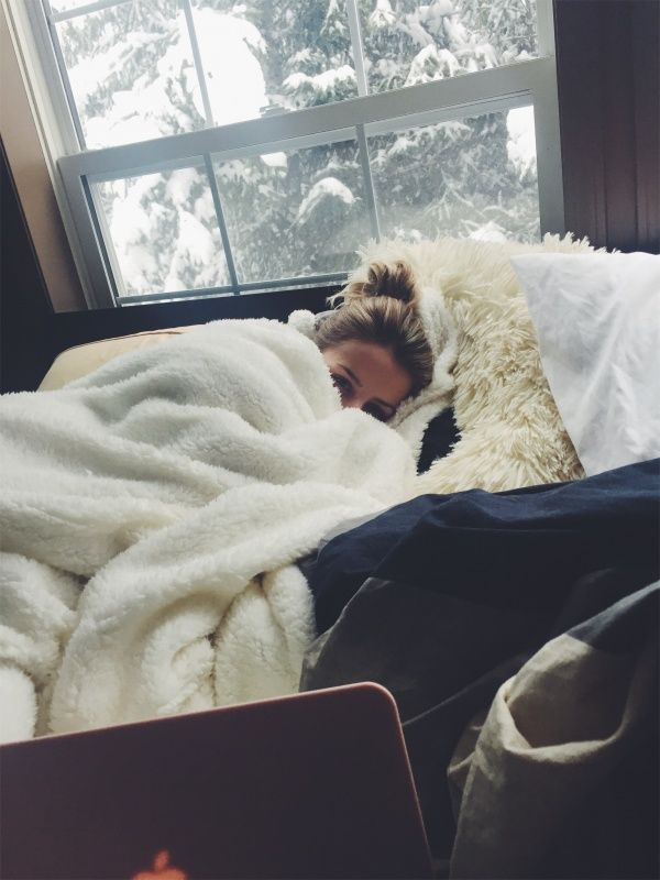 a woman laying in bed under a blanket next to a window with snow on it