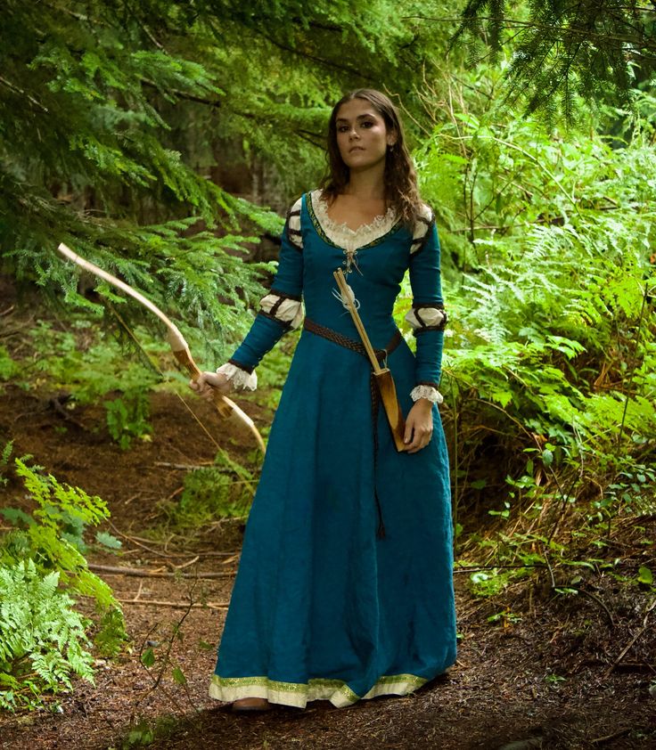 a woman in a blue dress holding a bow and arrow standing on a forest path