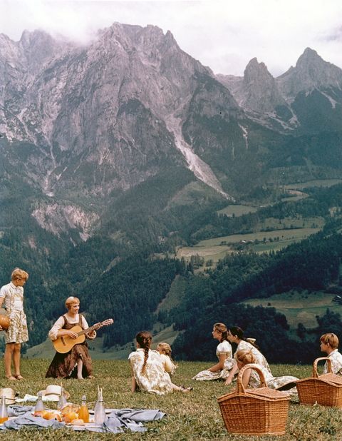 group of people sitting on the grass in front of mountains playing guitar and drinking wine