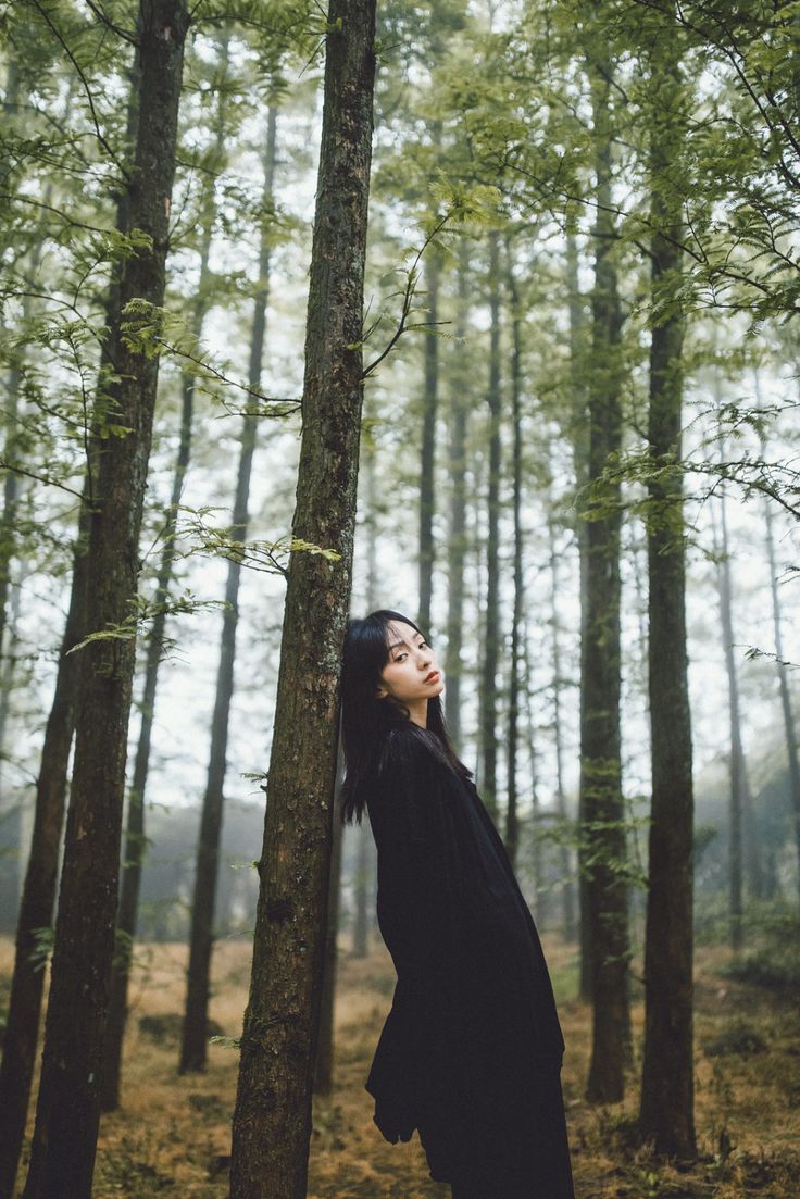 a woman standing in the middle of a forest with her head up against a tree