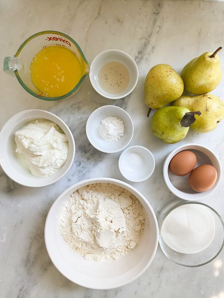 ingredients to make pear cake laid out on a marble counter top, including eggs, flour, butter and pears