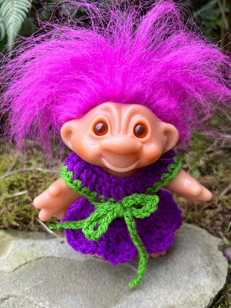 a troll doll with purple hair and green pants on top of a rock in the grass