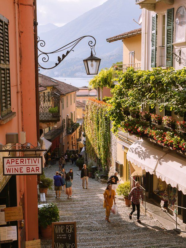 people are walking down an alley way in the mountainside town with flowers on the balconies