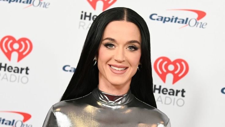 a woman with long black hair wearing a metallic outfit and smiling at the camera while standing in front of a heart radio logo