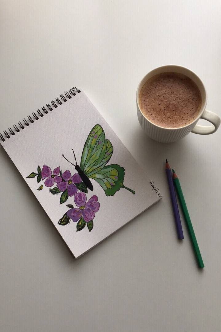 a cup of coffee next to a notebook with a butterfly on it and two colored pencils