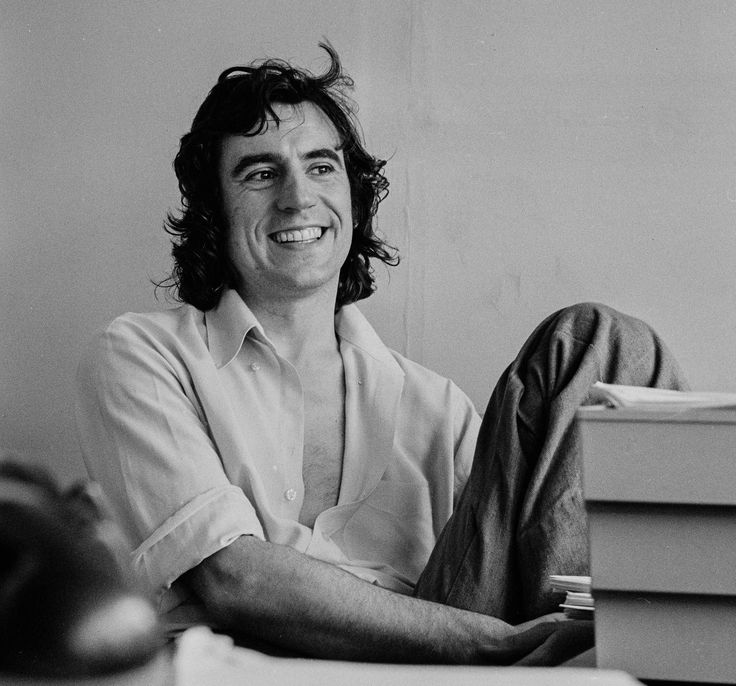 a black and white photo of a man sitting on a desk smiling at the camera