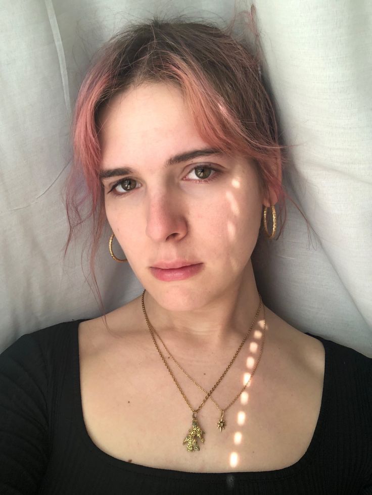 a woman with pink hair laying in bed wearing a black shirt and gold necklaces