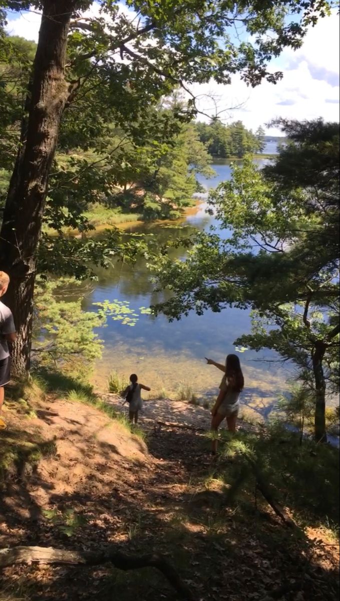 two people standing on the side of a hill next to a body of water and trees