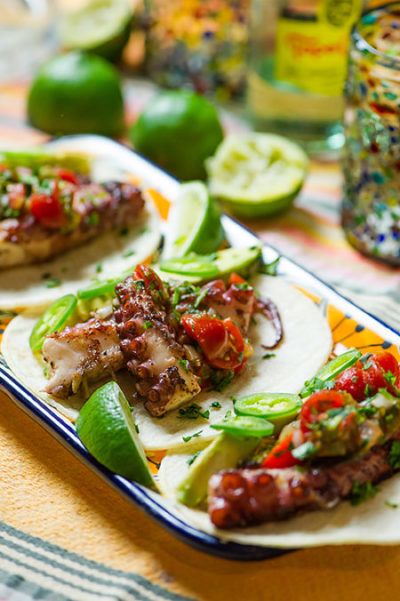 three tacos on a plate with limes, tomatoes and other ingredients around them