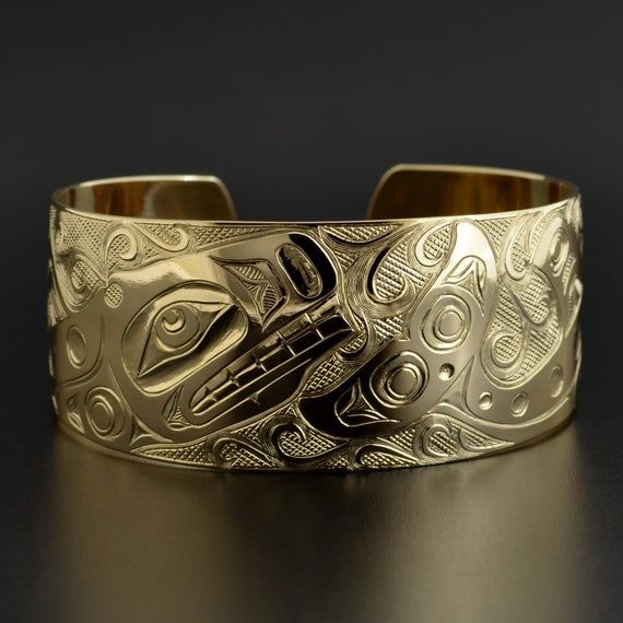 This cuff is hand-engraved and measures 1" wide by 6" long and was designed by First Nations artist Kelvin Thompson. This will fit a wrist between 6 1/4" and 6 1/2" in circumference. Custom lengths can be made on request. This piece is made to order and is titled 'Pod of Whales'.Whales usually signify longevity and strong community bonds due to their tendency to travel in close multi-family pods. In artworks, Orcas can be identified by a large, snub-nosed head; elongated nostrils; a wide, toothy Carved Symbolic Yellow Gold Jewelry, Yellow Gold Symbolic Carved Jewelry, Ceremonial Gold Bracelets With Polished Finish, Wide Band Yellow Gold Bracelets As Gift, Wide Band Yellow Gold Bracelets For Gift, Unique Yellow Gold Cuff Bracelet For Formal Occasions, Luxury Engraved Round Cuff Bracelet, Luxury Engraved Cuff Bracelet For Anniversary, Formal Engraved Bangle Cuff Bracelet