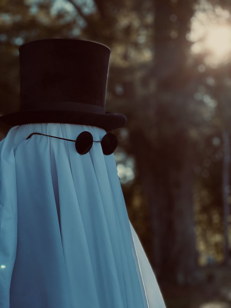 a close up of a person wearing a white dress and black top hat with trees in the background