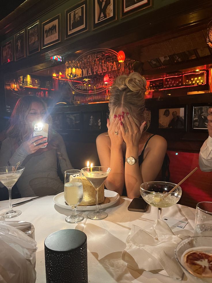 two women sitting at a table with food and drinks in front of them, one holding her face