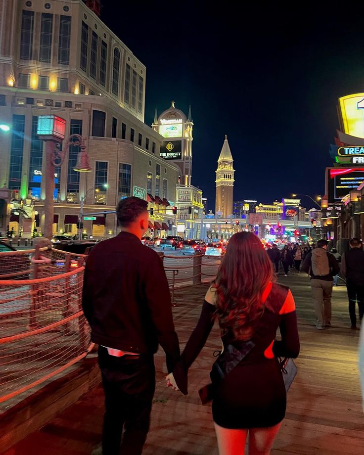 a man and woman holding hands walking down the street in front of some buildings at night