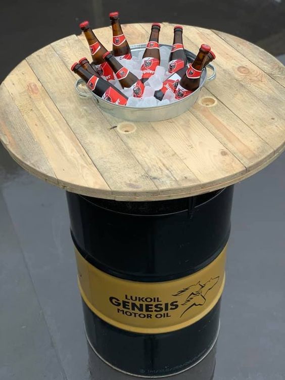 a wooden table topped with bottles of beer on top of a black barrel filled with ice