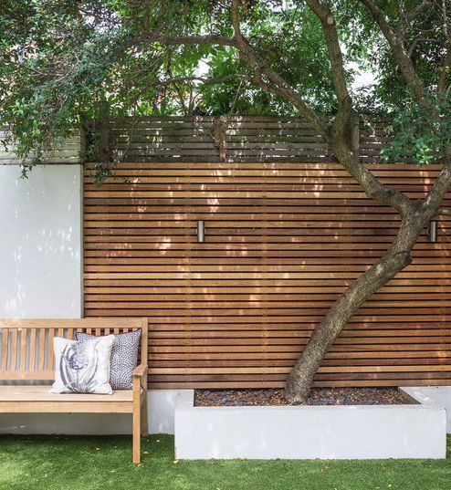 a wooden bench sitting under a tree next to a white wall and green grass covered ground