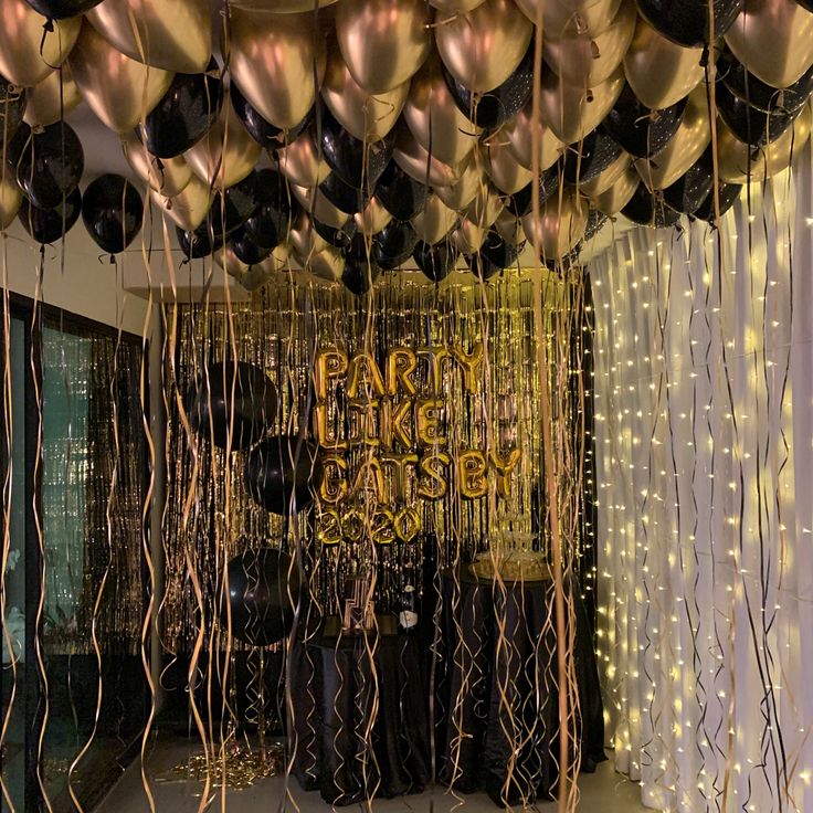 balloons and streamers are hanging from the ceiling in front of a party backdrop with black and gold decorations