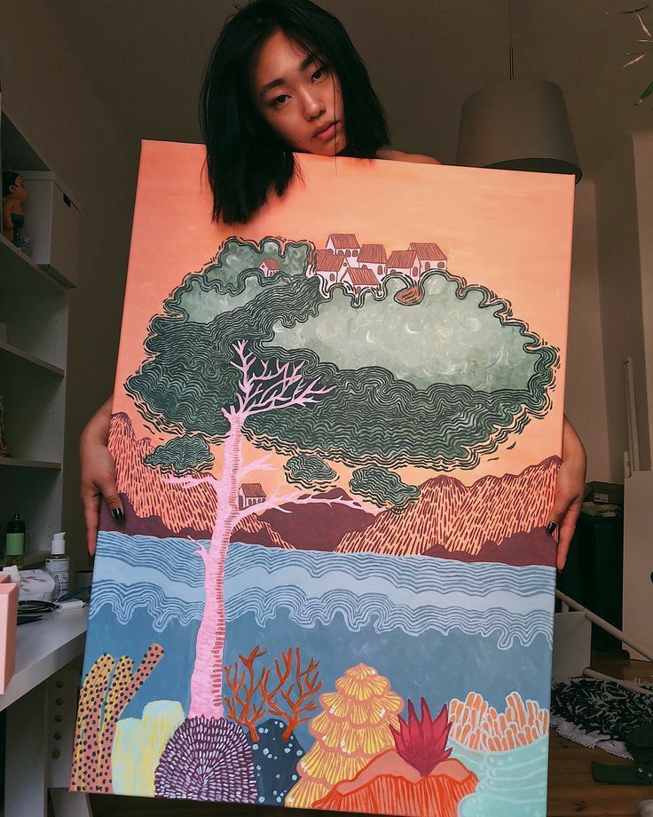 a woman holding up an art piece in front of her face with trees and mountains painted on it