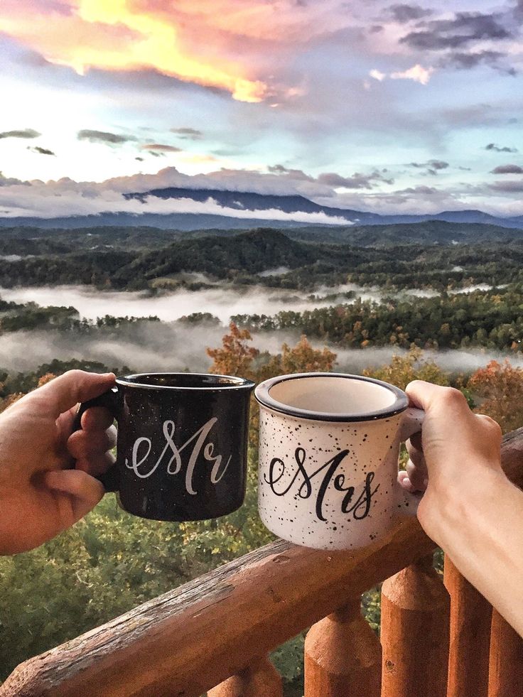 two people holding coffee mugs on top of a wooden railing overlooking a valley and mountains