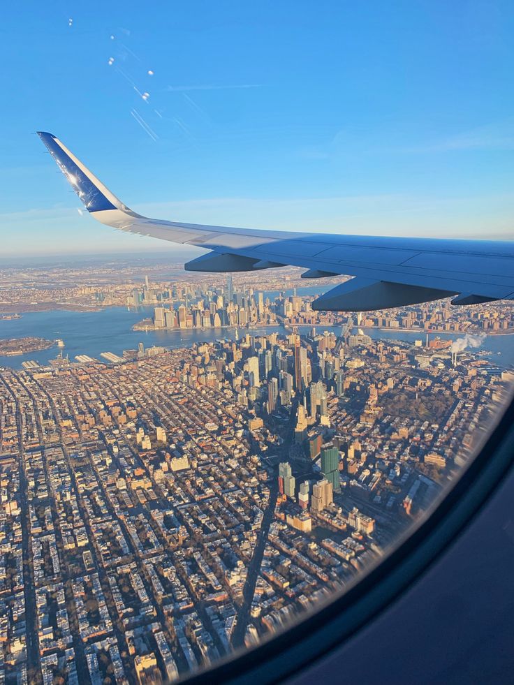 an airplane wing flying over a city with tall buildings in the background and water below