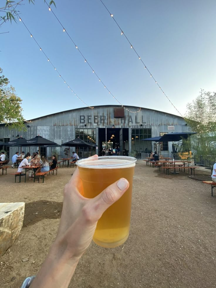 a person holding up a beer glass in front of a building with tables and umbrellas