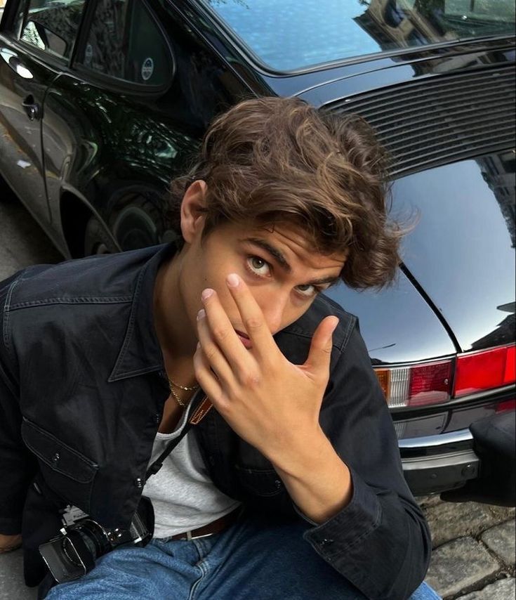 a young man sitting next to a parked car holding his hand up to his mouth