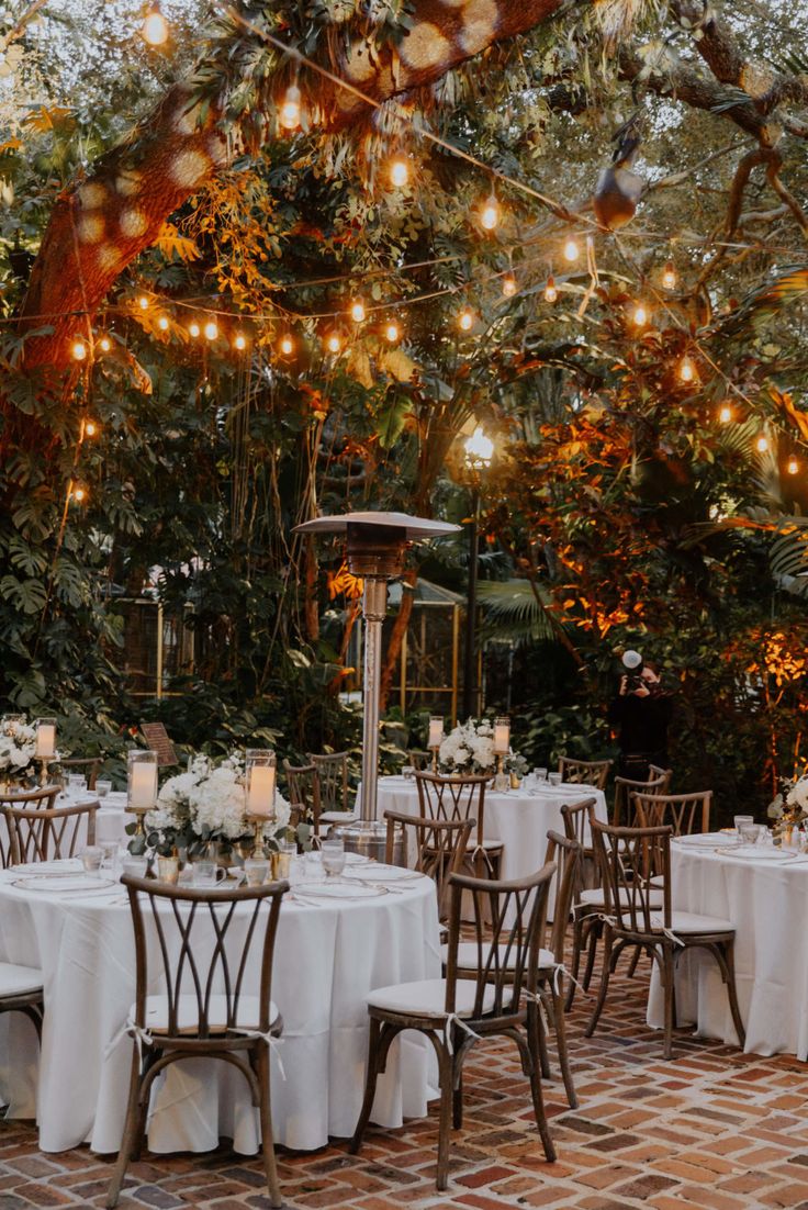 an outdoor wedding reception with white linens and lights hanging from the ceiling, surrounded by greenery