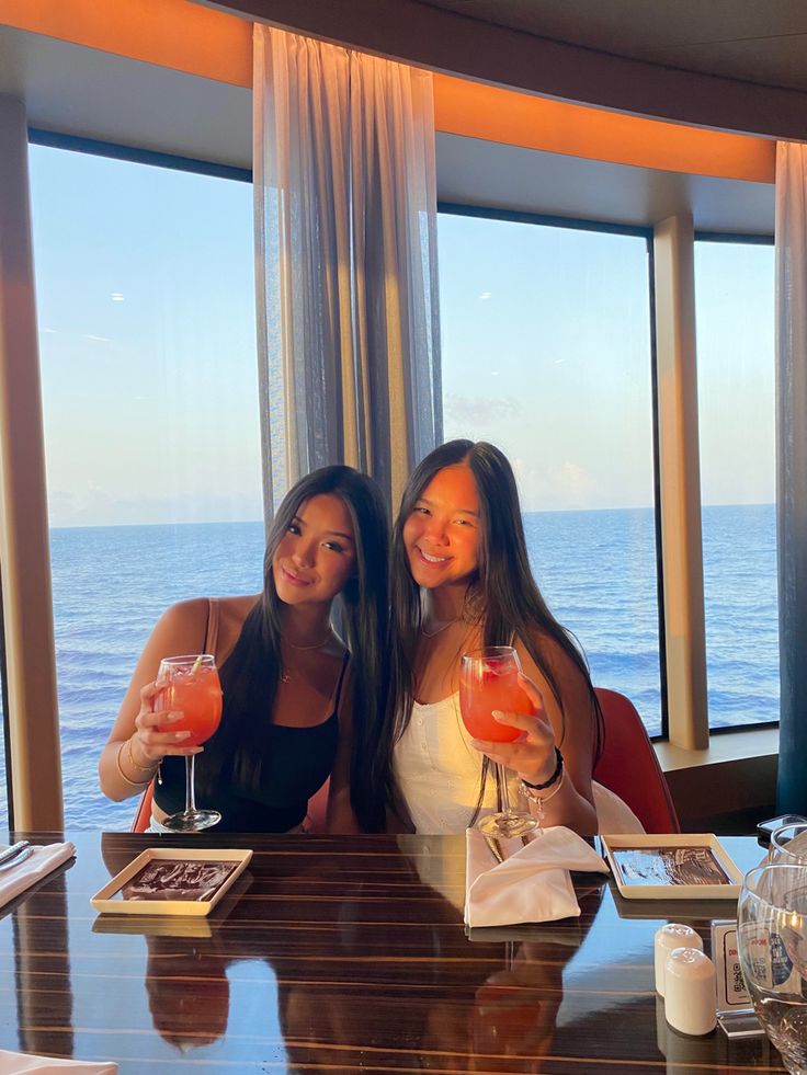 two beautiful young women sitting at a table with drinks in front of large windows overlooking the ocean