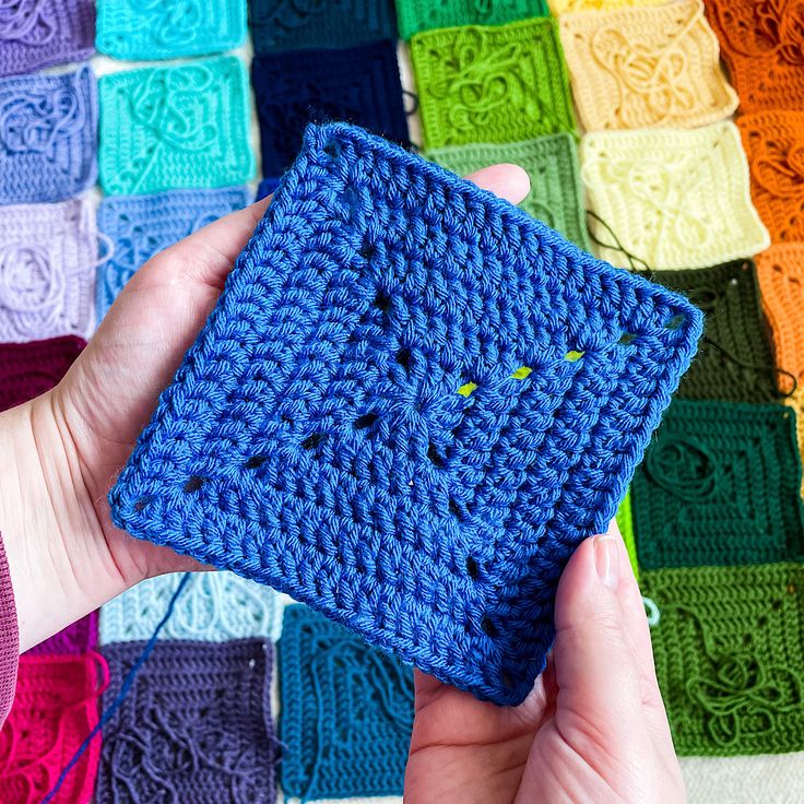 a person holding up a crocheted square in front of a multicolored blanket