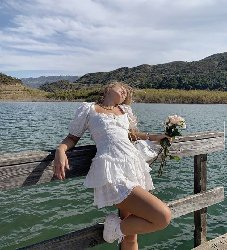 a beautiful young woman leaning on a wooden fence next to the water holding a bouquet of flowers