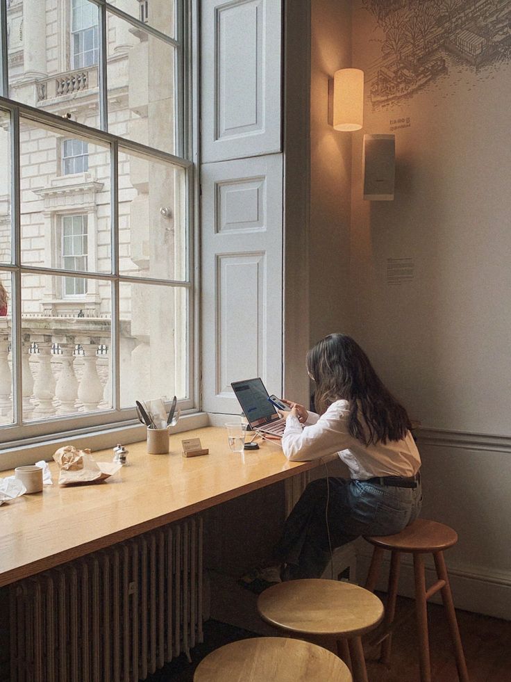 a woman sitting at a table with a laptop computer in front of a large window