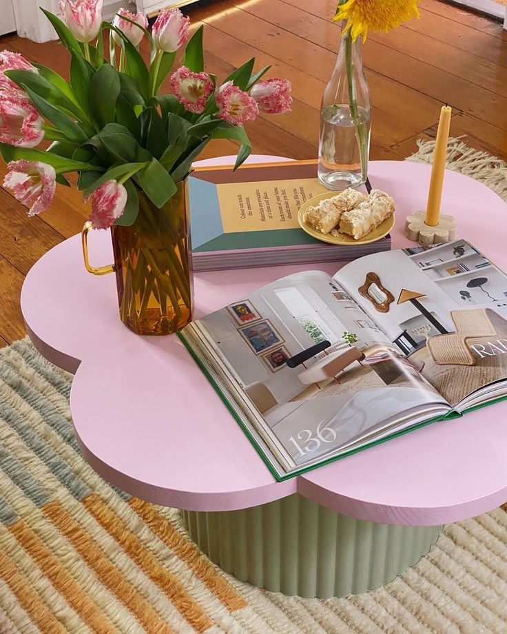 a pink coffee table with magazines and flowers on it in front of a glass vase