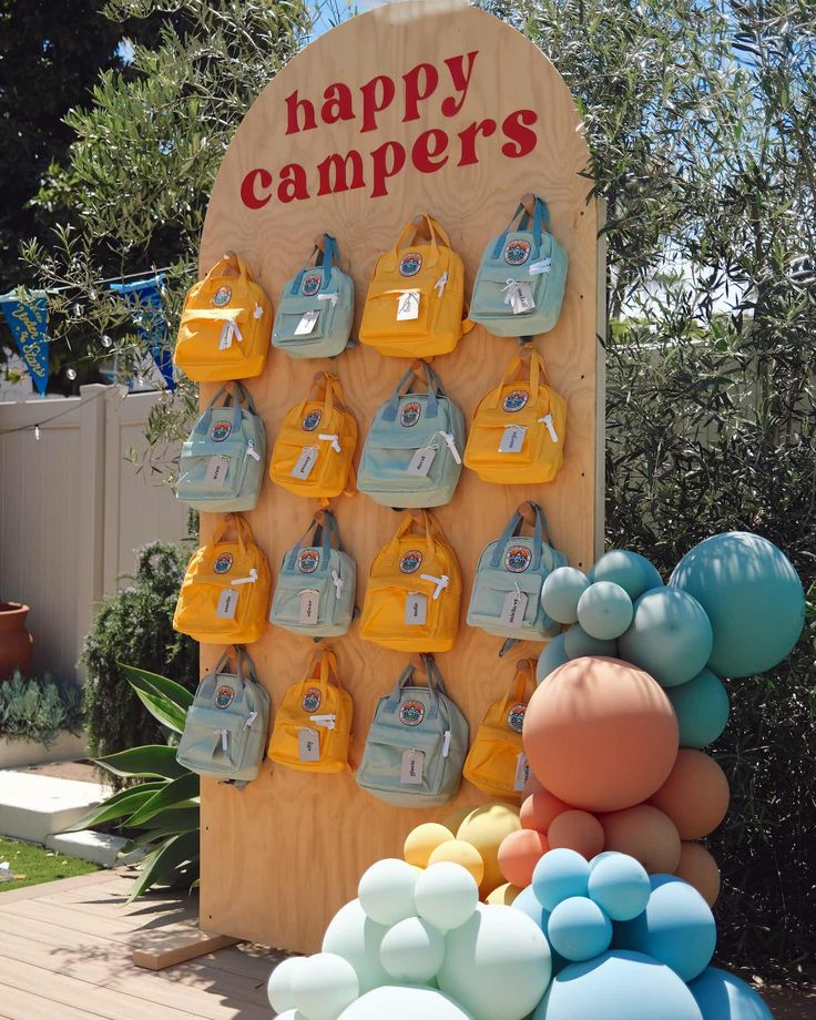 A large wooden slab with signage atop that says "Happy Campers" in red letters with yellow and blue backpacks in multiple mini sizes hanging from it and various balloons in size and color at the base Explorer Birthday Theme, Camping One Year Birthday, Campground Birthday Party, Camp Party Decor, Indoor Camping Birthday Party, Camp Birthday Theme, Camp Theme Birthday Party, Camp Party Theme, Camping First Birthday Party