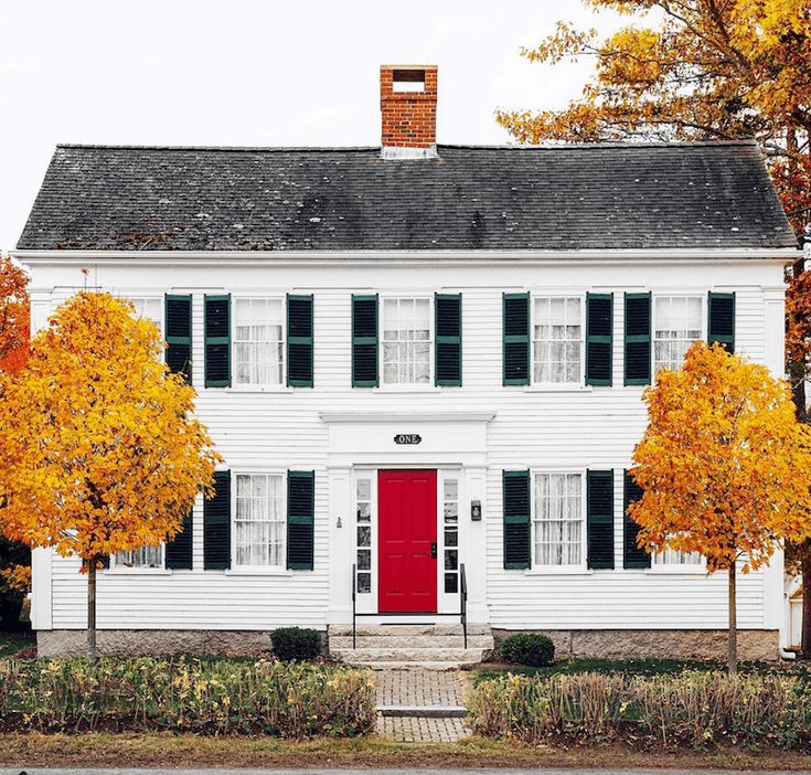 an old white house with red door and black shutters on the front, surrounded by autumn trees