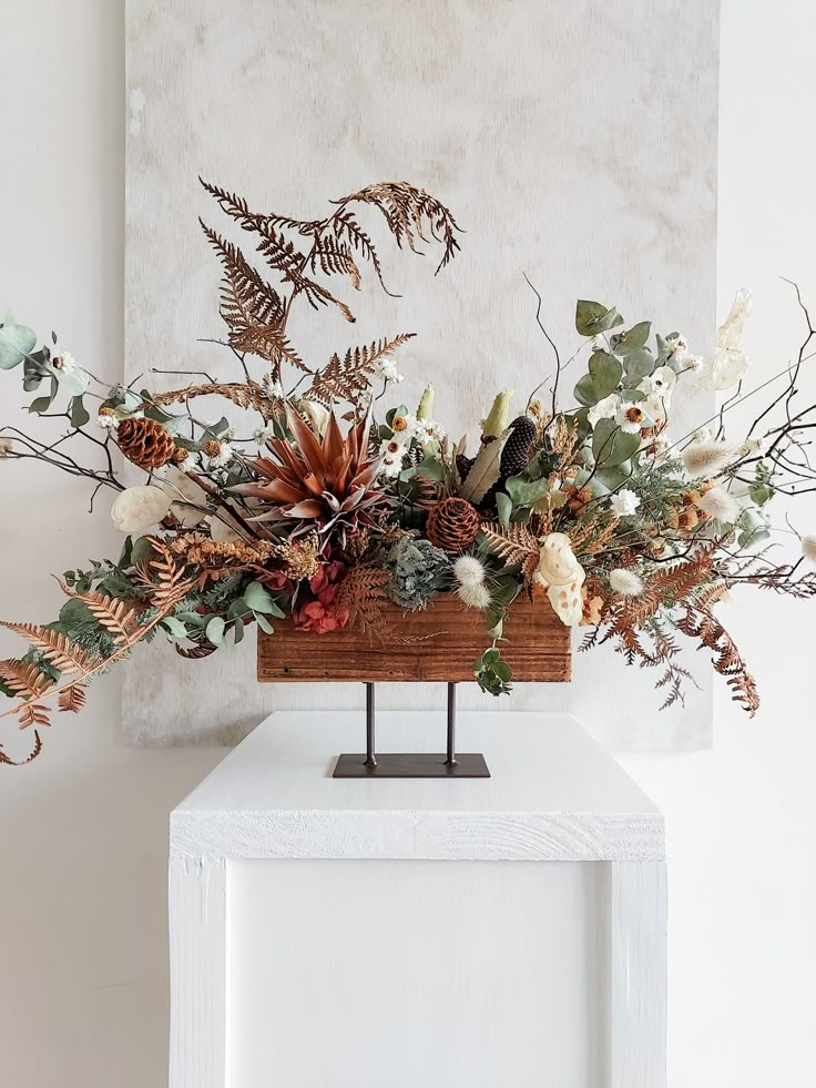 an arrangement of flowers and foliage on a white table in front of a wall hanging