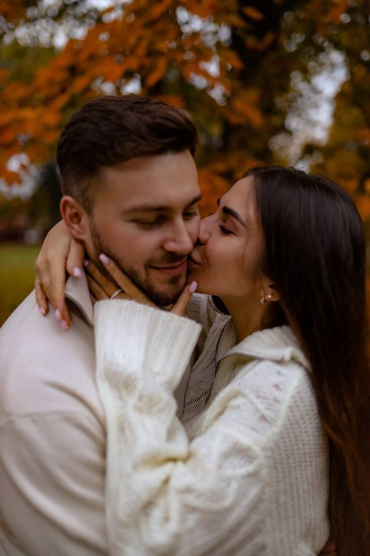 a man and woman kissing in front of trees with fall leaves on the branches behind them