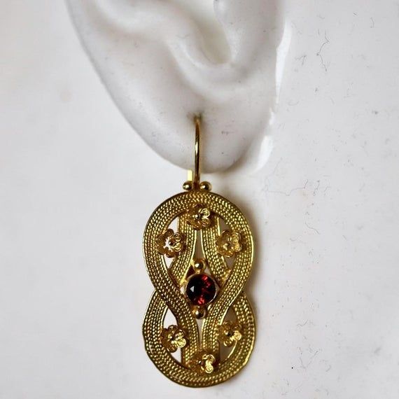 Hellenistic Greek motifs such as this Heracles knot design dating from the fourth century B.C. remained popular well into Roman times. Handcrafted of filigree wire accented with rosettes, it is inlaid with a faceted garnet. 14k gold settings, with European back for pierced ears. Size 1 1/2 Inches. Historical Yellow Gold Drop Earrings, Yellow Gold Drop Earrings With Historical Design, Yellow Gold Historical Drop Earrings, Traditional Formal Jewelry With Historical Design, Elegant Oval Earrings With Intricate Design, Victorian Filigree Oval Earrings, Victorian Oval Filigree Earrings, Yellow Gold Earrings With Historical Design As Gift, Elegant Ceremonial Earrings