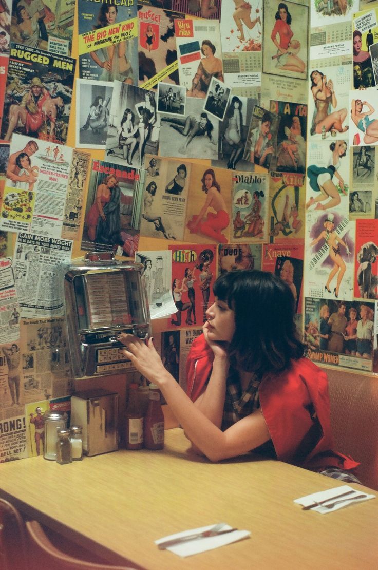 a woman sitting at a table in front of a wall covered with old advertisements and posters