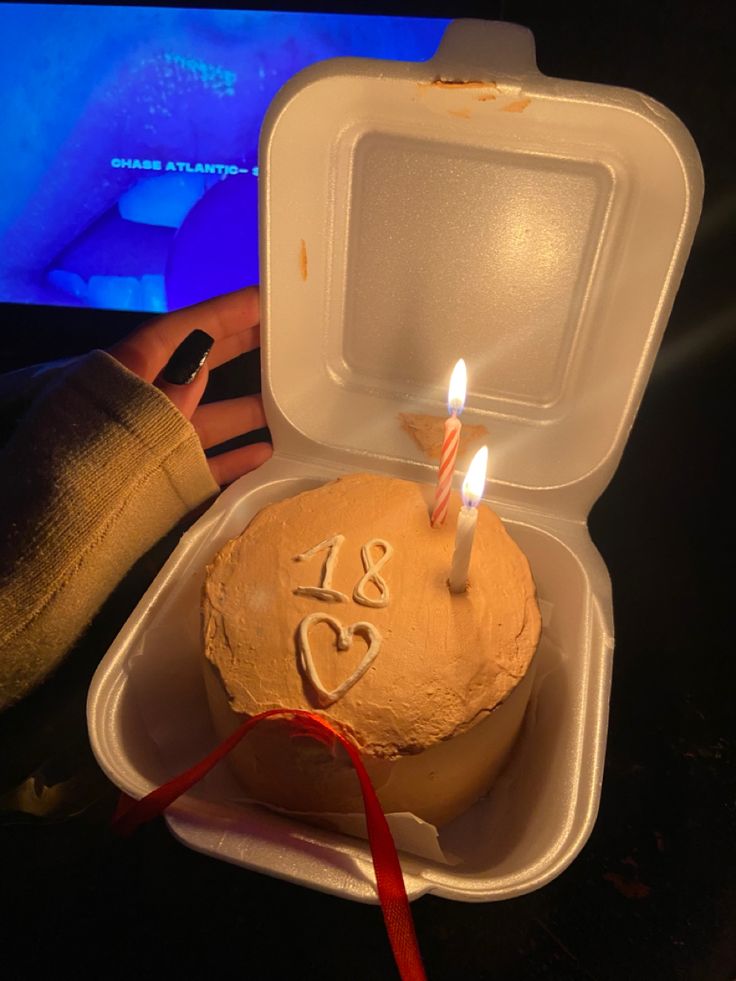 a birthday cake in a foam container with two lit candles on the top, and someone's hand next to it