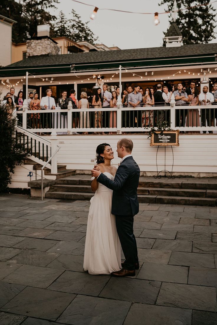 a bride and groom dance together in front of an outdoor venue with people watching from the balcony