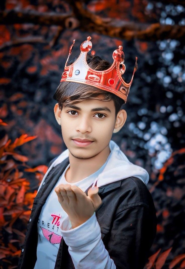 a young boy wearing a red crown standing in front of some trees and leaves with his hand on his chest