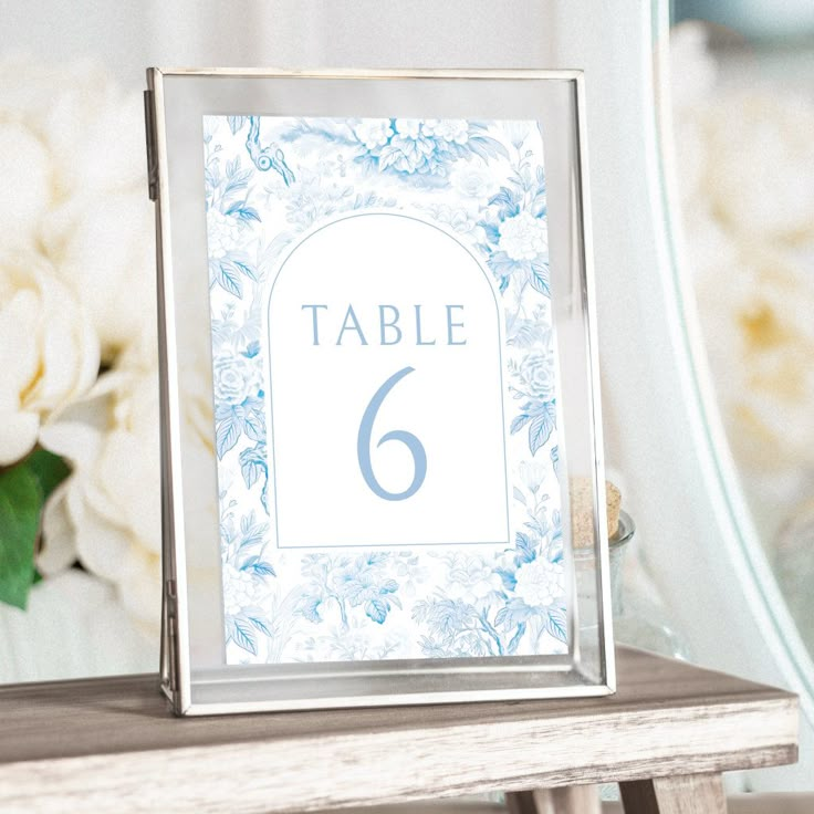 a table number sign sitting on top of a wooden table next to a vase with white flowers