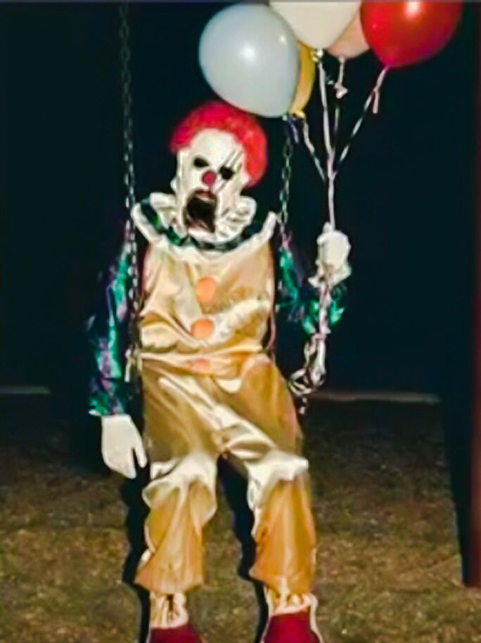 the clown is holding balloons in his hand and wearing red shoes, while sitting on a swing