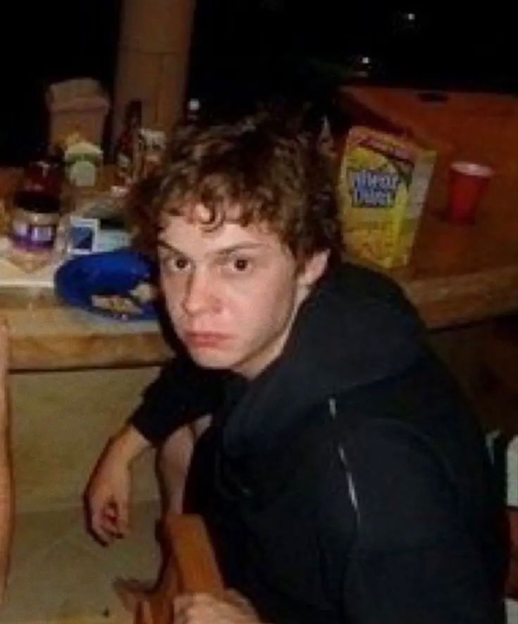 two young men sitting at a table with food on the counter and one is looking into the camera